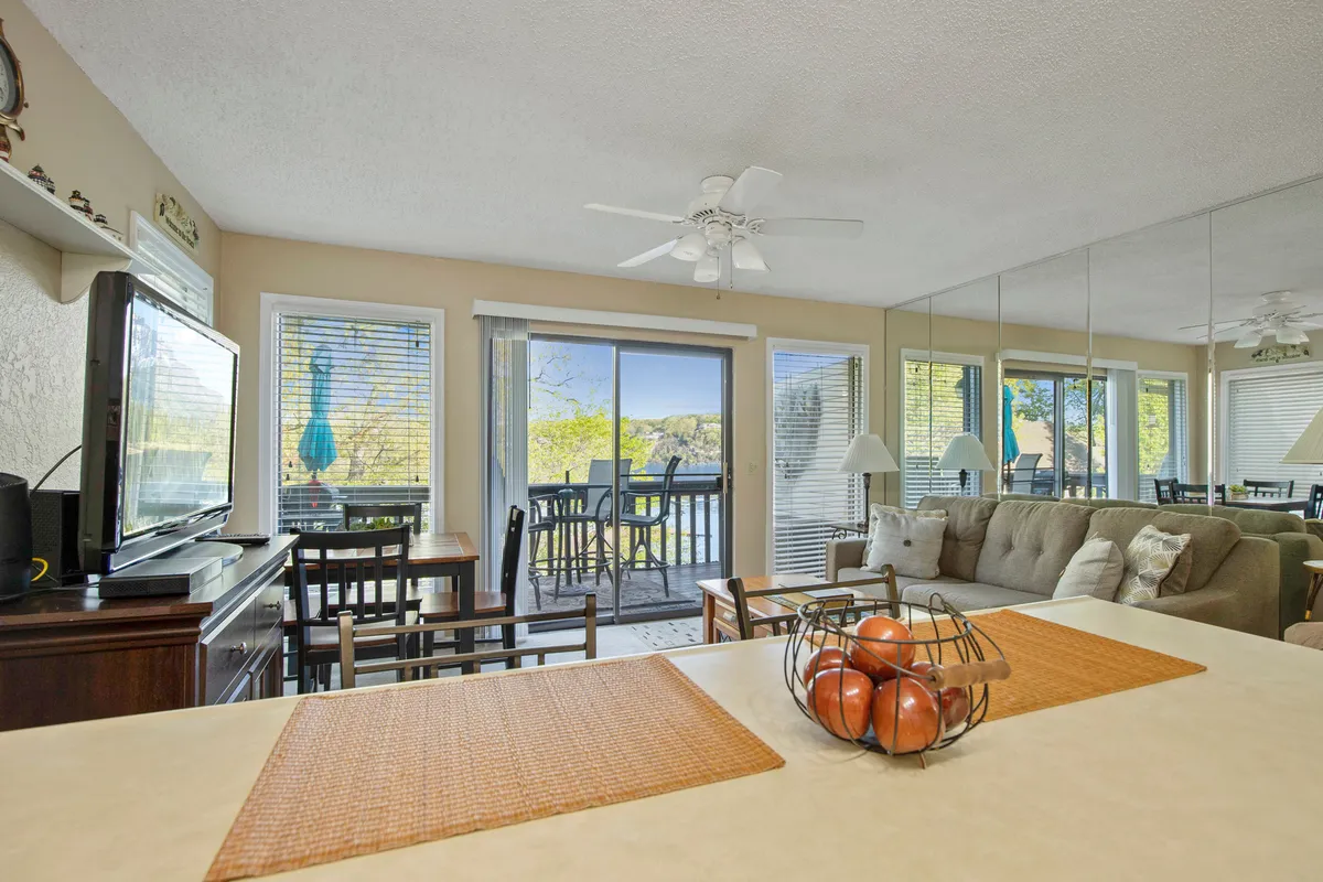 This image captures a bright, open living and dining area with a comfortable couch, a dining table with chairs, and a kitchen countertop adorned with a fruit bowl. Large windows fill the space with natural light and offer an inviting view of the outdoors, reinforcing the airy, lakehouse atmosphere.