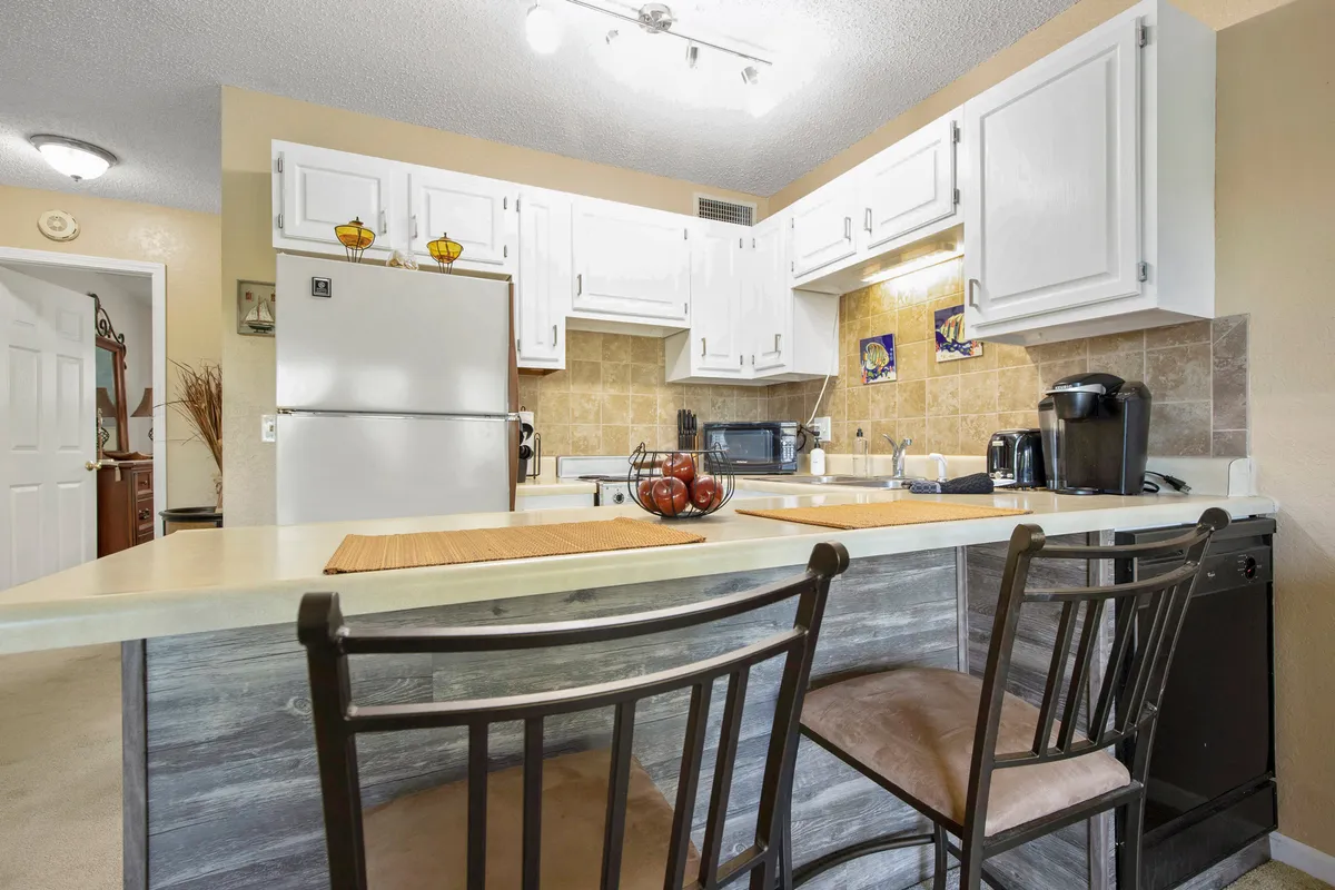 A bright, welcoming kitchen featuring a spacious island with bar stools, white cabinetry, and modern appliances. The setup suggests ample workspace for meal prep and invites socializing during cooking. Small touches, like a coffee maker and neatly arranged essentials, hint at a well-stocked kitchen ready for both gourmet creations and casual meals.
