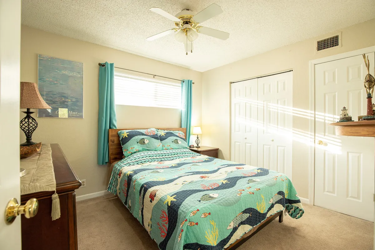 A cozy bedroom featuring a bed adorned with a colorful quilt, a nightstand with a lamp, and a decorative painting on the wall. The room is bathed in warm, relaxing sunlight, and the décor is calm and inviting.