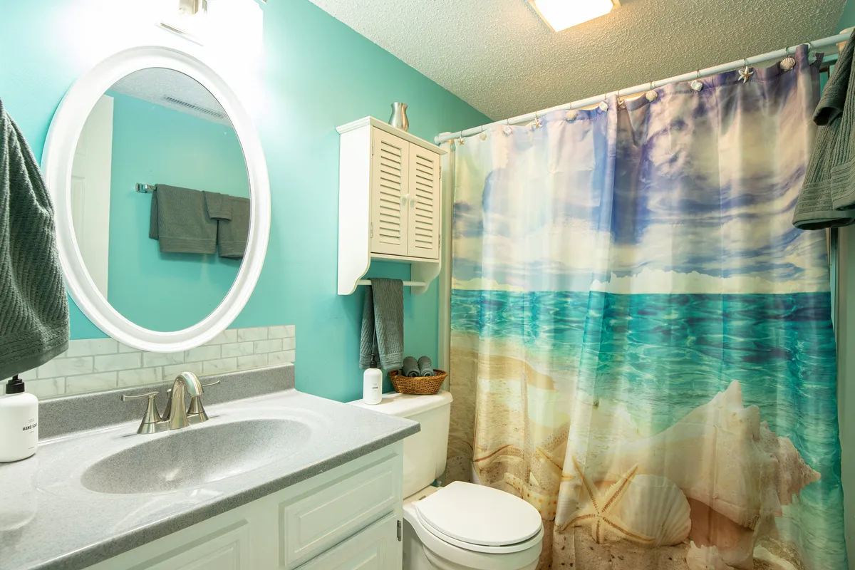A bright, serene bathroom featuring a large round mirror, a beach-themed shower curtain, and light blue walls. The space feels airy and clean, with subtle touches that evoke a spa-like ambiance.