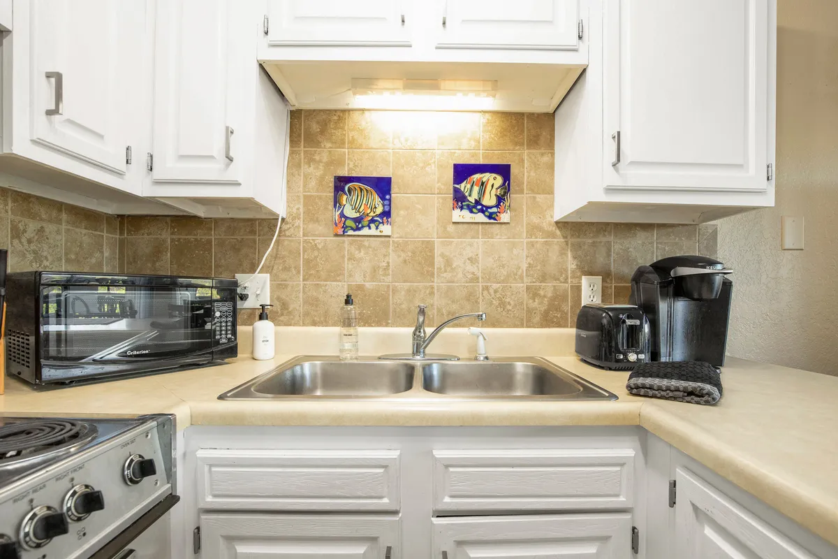 A cheerful kitchen vignette with a focus on the sink area, decorative fish paintings, a microwave, and a prominent coffee maker. The overall mood is bright and functional, with homey details suggesting comfort and readiness for daily routines or special breakfasts.